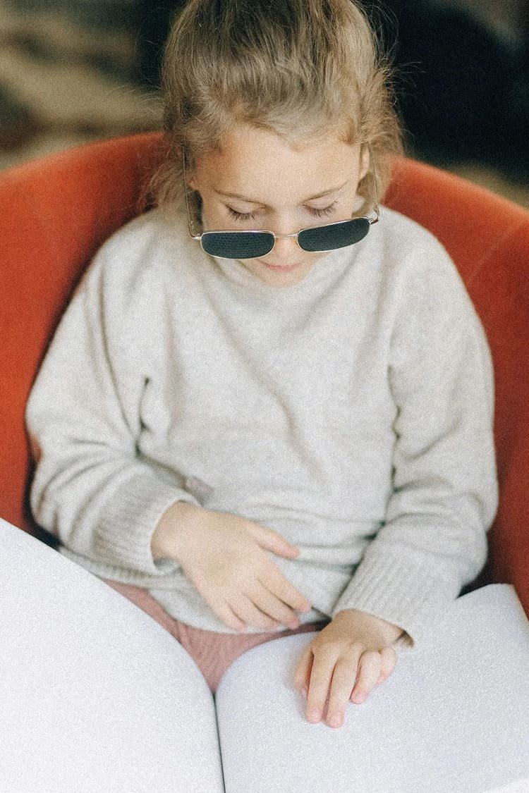 A young girl with light brown hair wearing sunglasses, a light gray sweater, sitting in a red chair, looking down at a book.