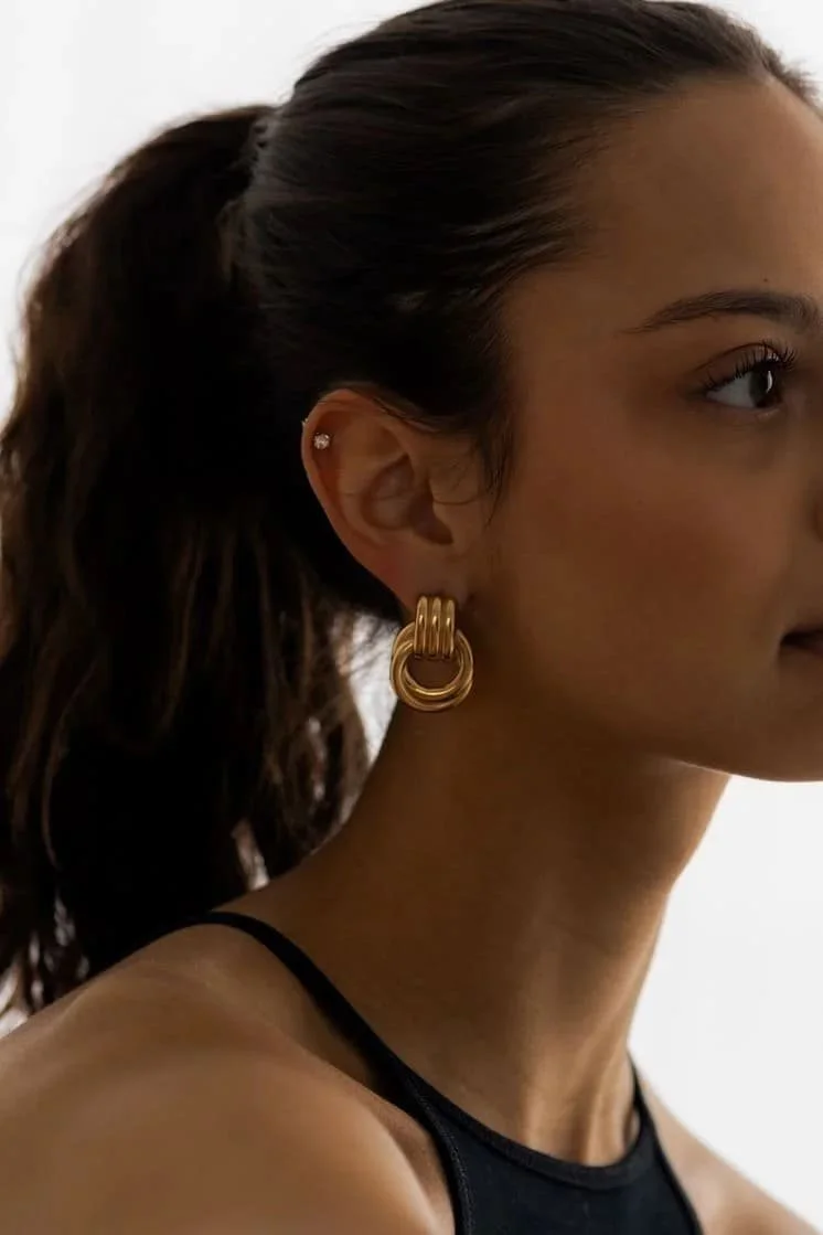 A woman with brown hair tied in a ponytail wearing gold hoop earrings and a black strap top.