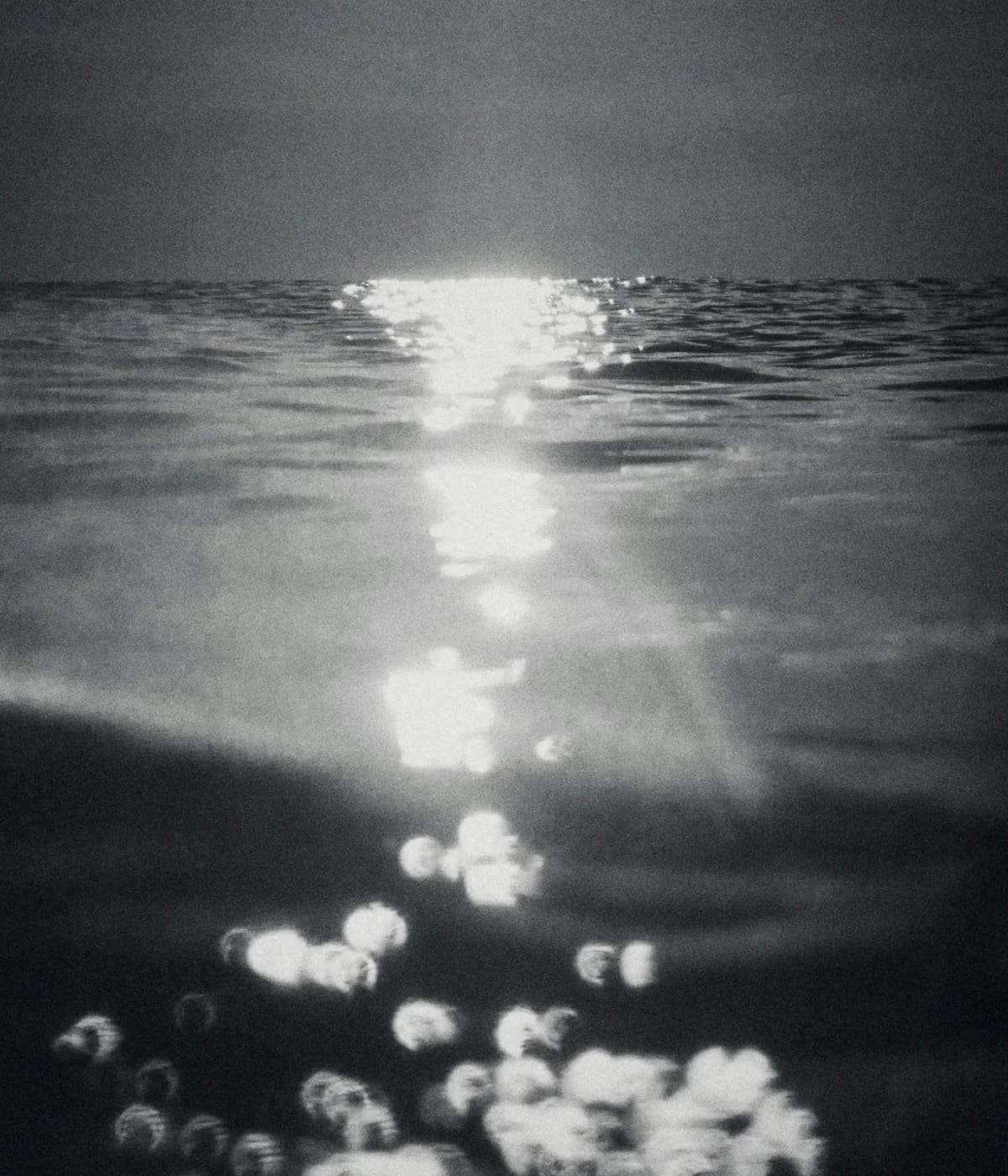 Black and white photo of the reflective surface of the ocean with sunlight creating a bright path on the water and the horizon line in the background.
