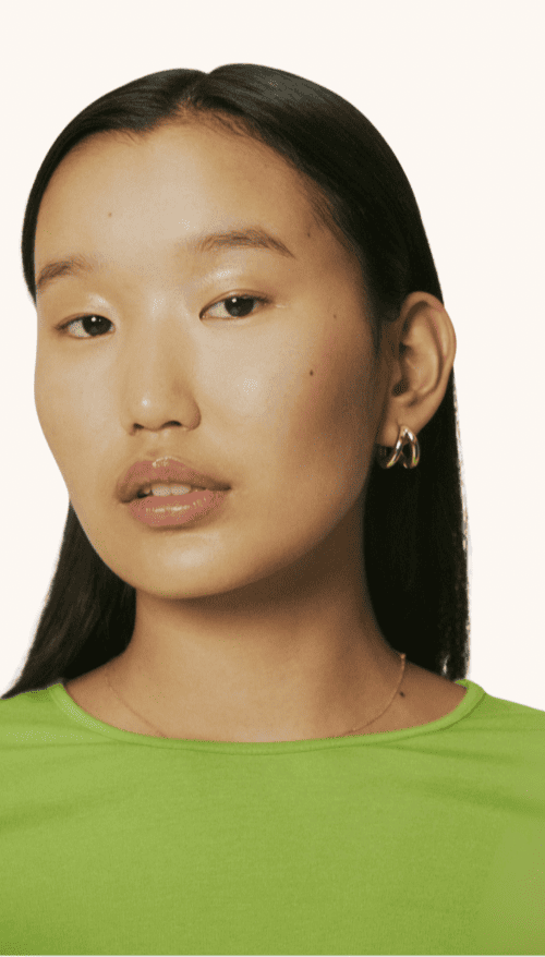 Close-up of a young Asian woman with long black hair, wearing silver hoop earrings and a bright green shirt, against a white background.