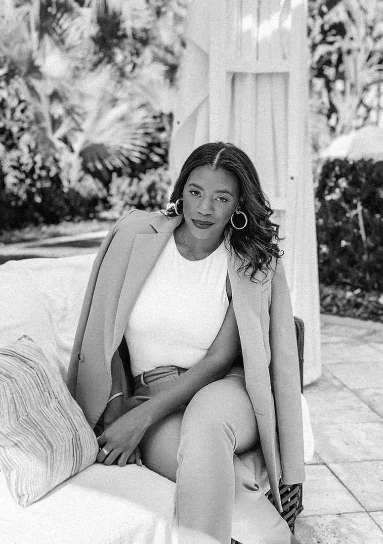A woman sitting on a sofa outdoors, wearing a light-colored blazer draped over her shoulders, a sleeveless top, and pants. She has medium-length curly hair, large hoop earrings, and is looking at the camera.