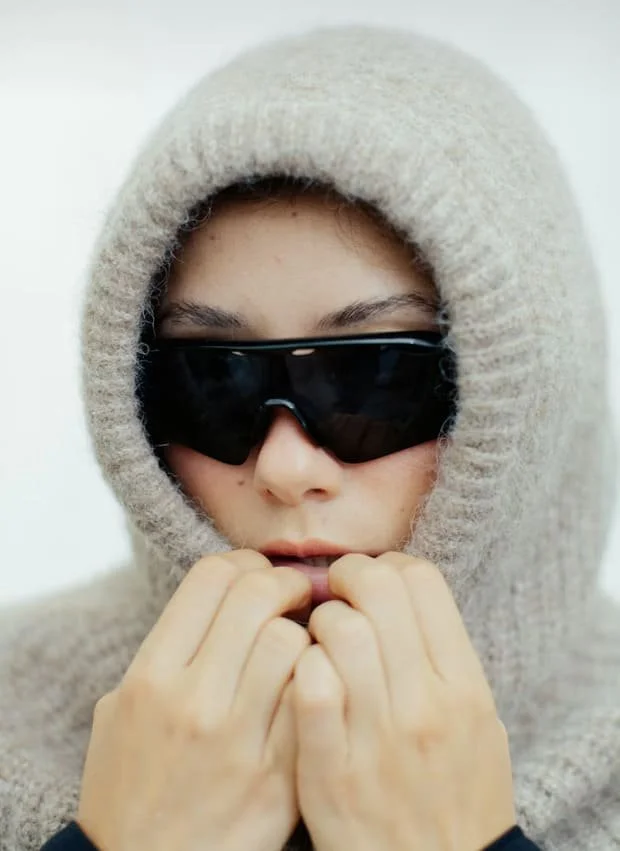 A person wearing a beige hoodie, black sunglasses, and holding their hands near their face, with a neutral background.