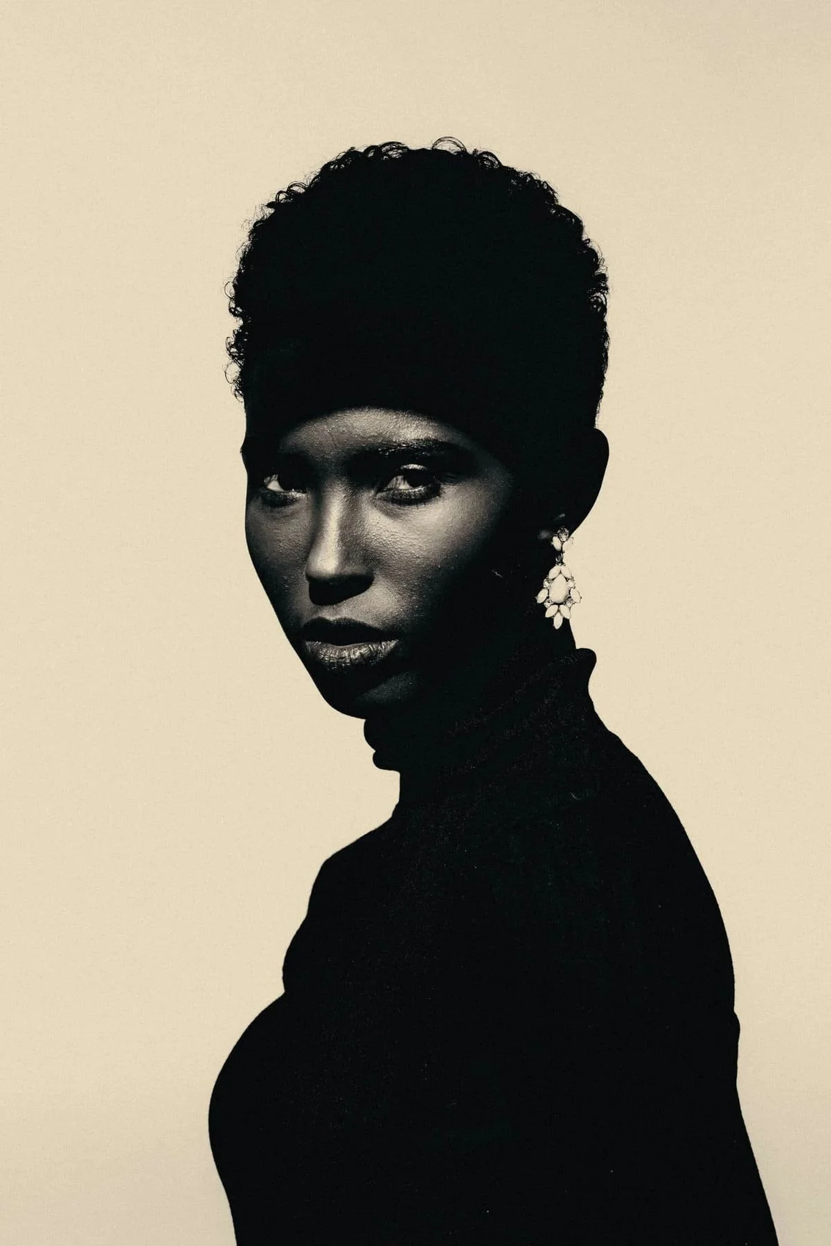 Black and white portrait of a woman with short curly hair, wearing earrings and a high-necked top, looking over her shoulder.