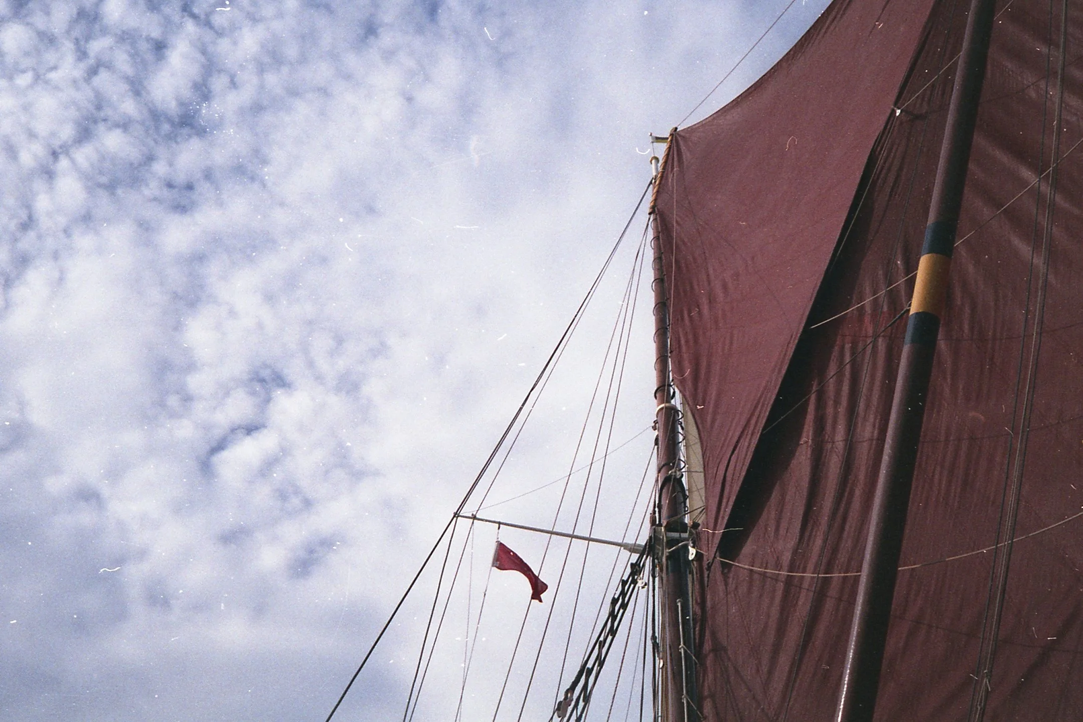 Close-up view looking up at the side of a sailboat with a large red sail and a small red flag, against a cloudy sky.