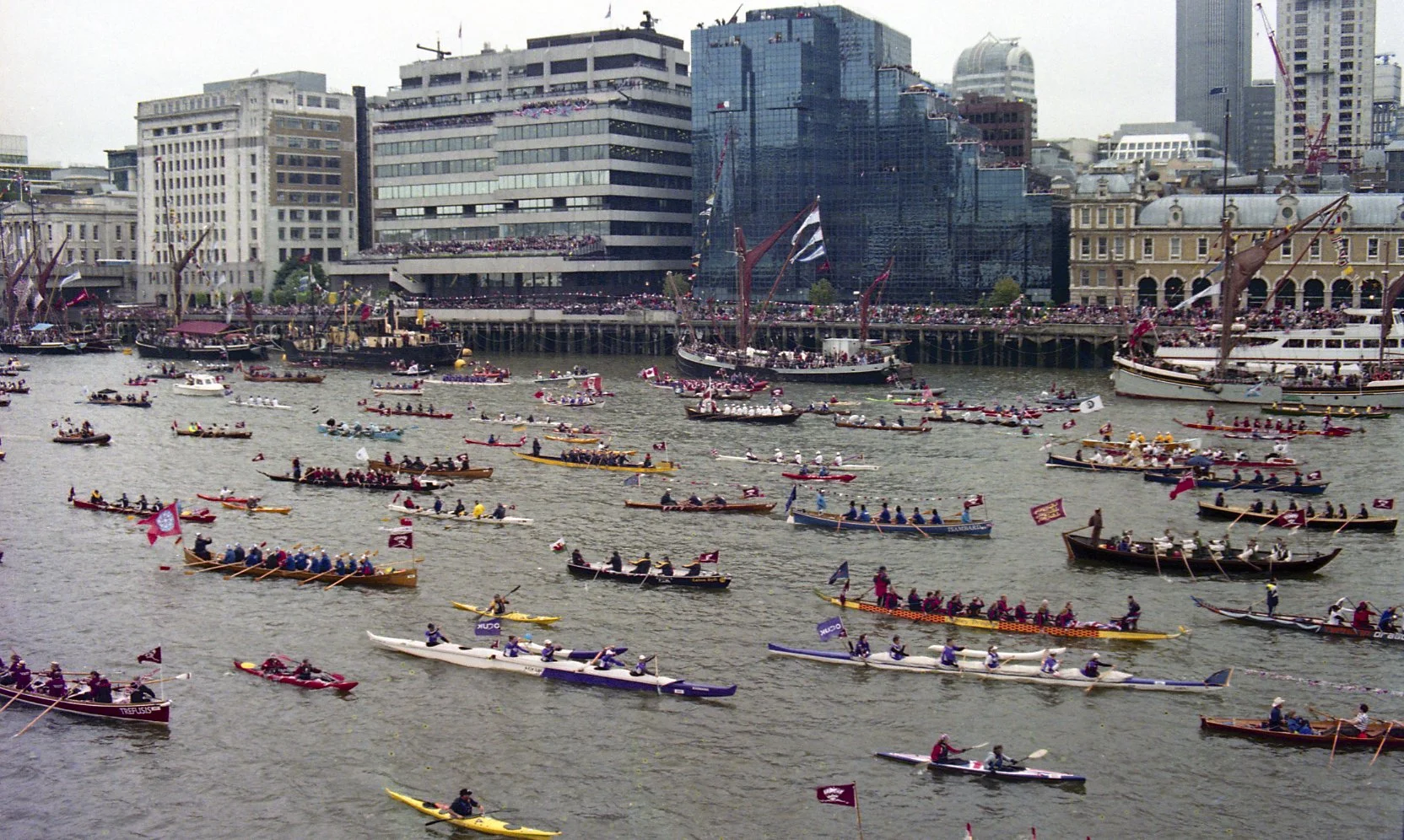 A large number of boats, many with flags, participate in a race on a river in an urban area, with tall buildings and a crowd of spectators on a pier.
