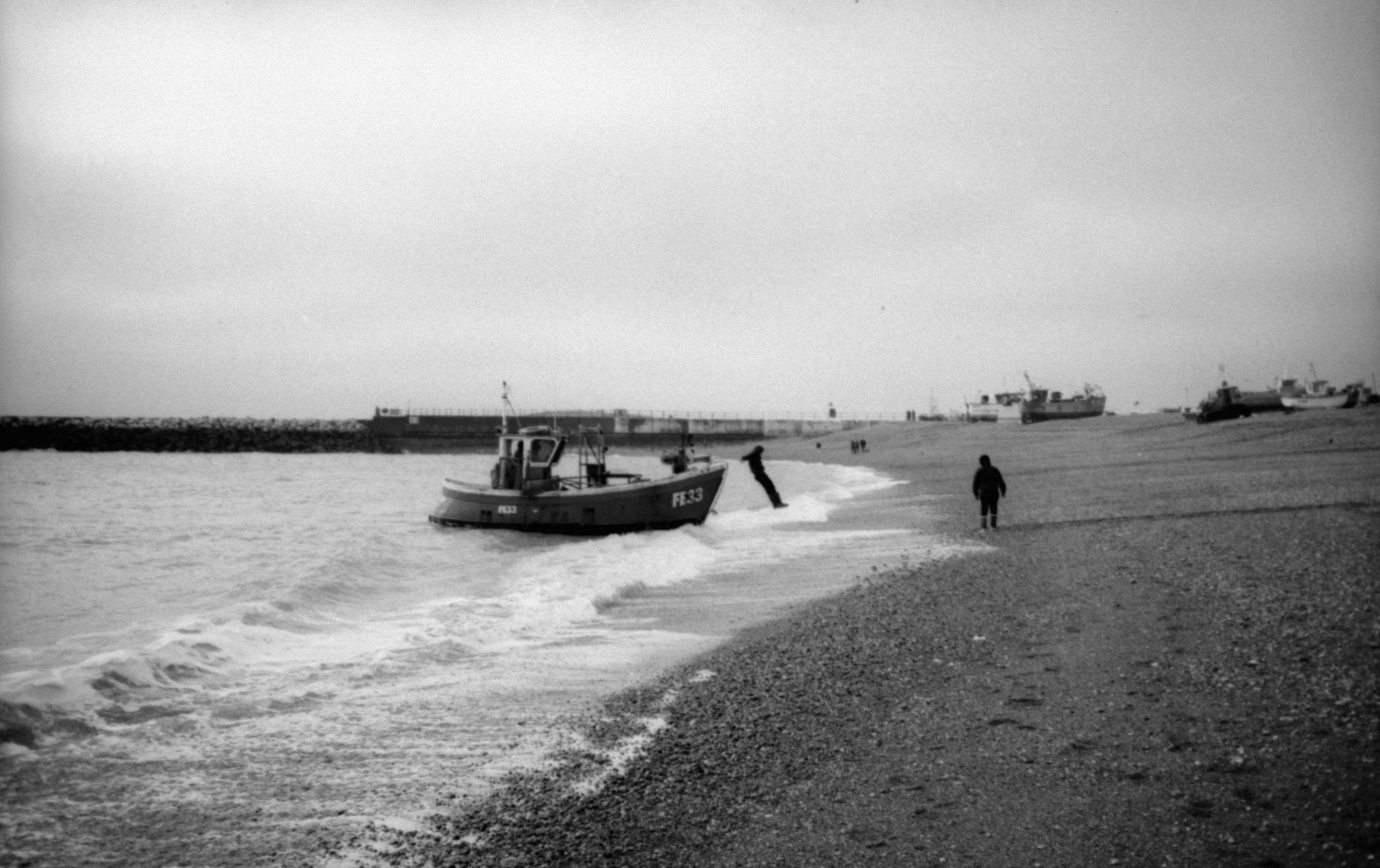 Black and white photograph of a beach scene with boats launched in the water and on the shore. Several people are walking along the beach. The sky appears overcast.