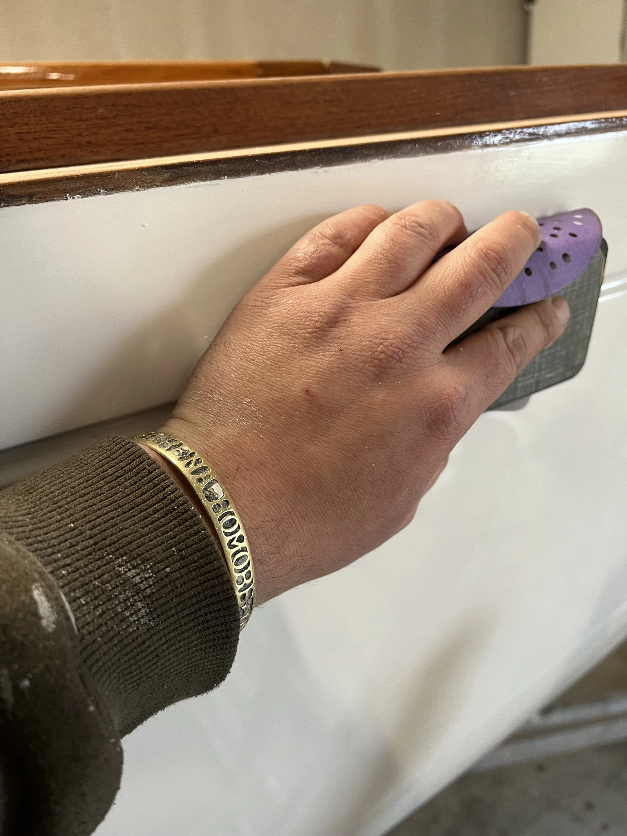 A person's hand holding a purple and black sanding tool against a white surface. The person is wearing a brown ribbed long sleeve shirt and a gold bracelet with black inscriptions.
