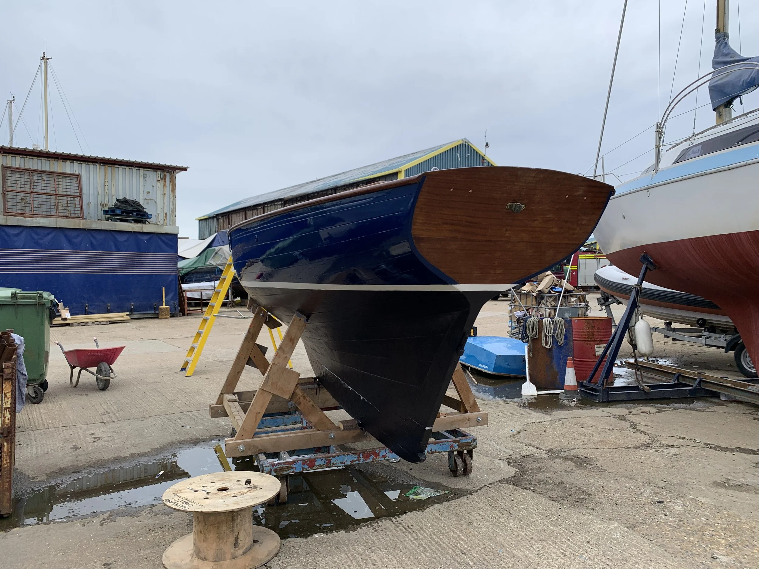 Boat on a wooden stand at a boatyard, with other boats, tools, and a shed in the background.