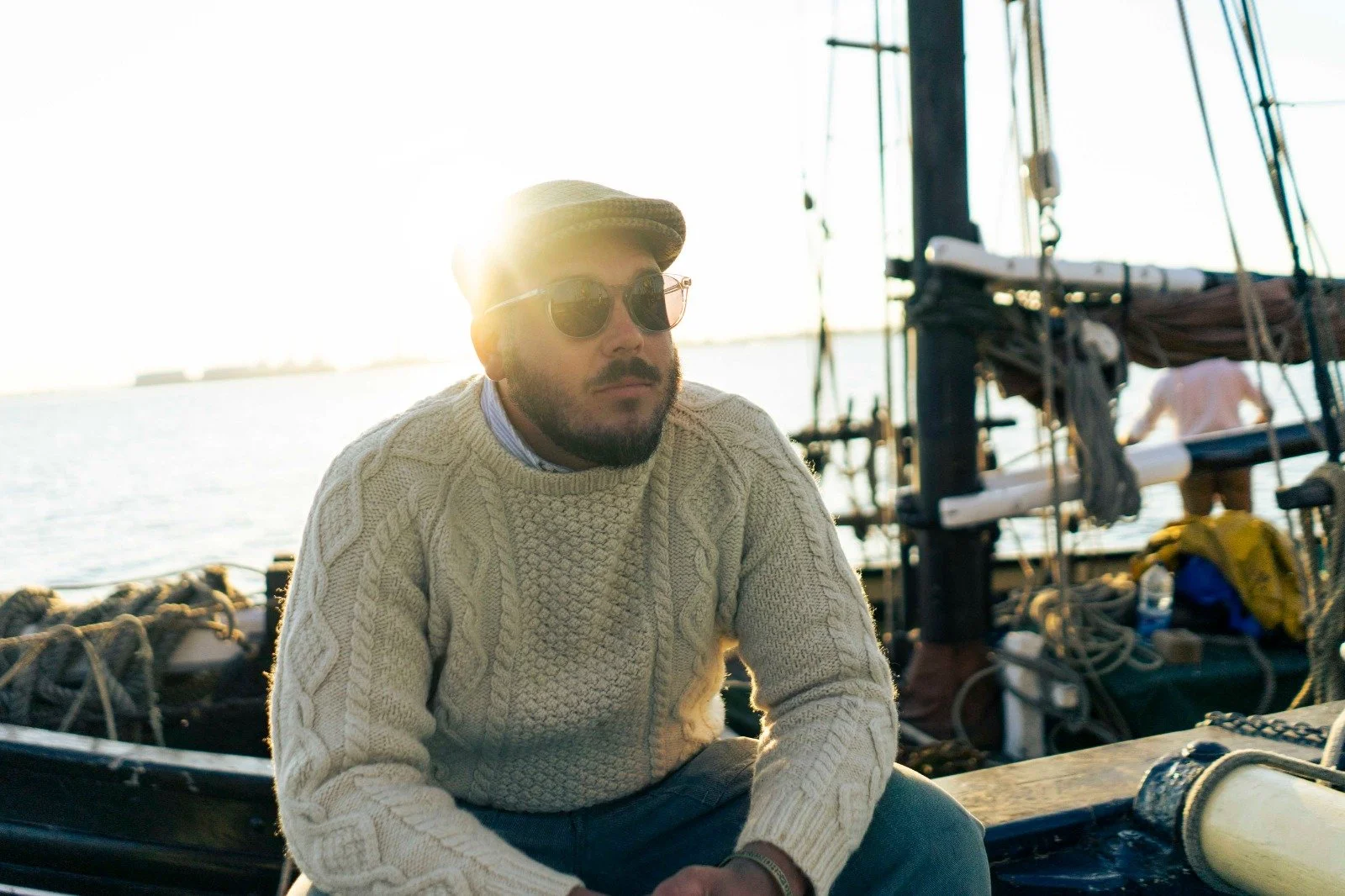 A man in a cream-colored cable knit sweater, wearing sunglasses and a flat cap, sitting on a boat with water and sailing equipment in the background.