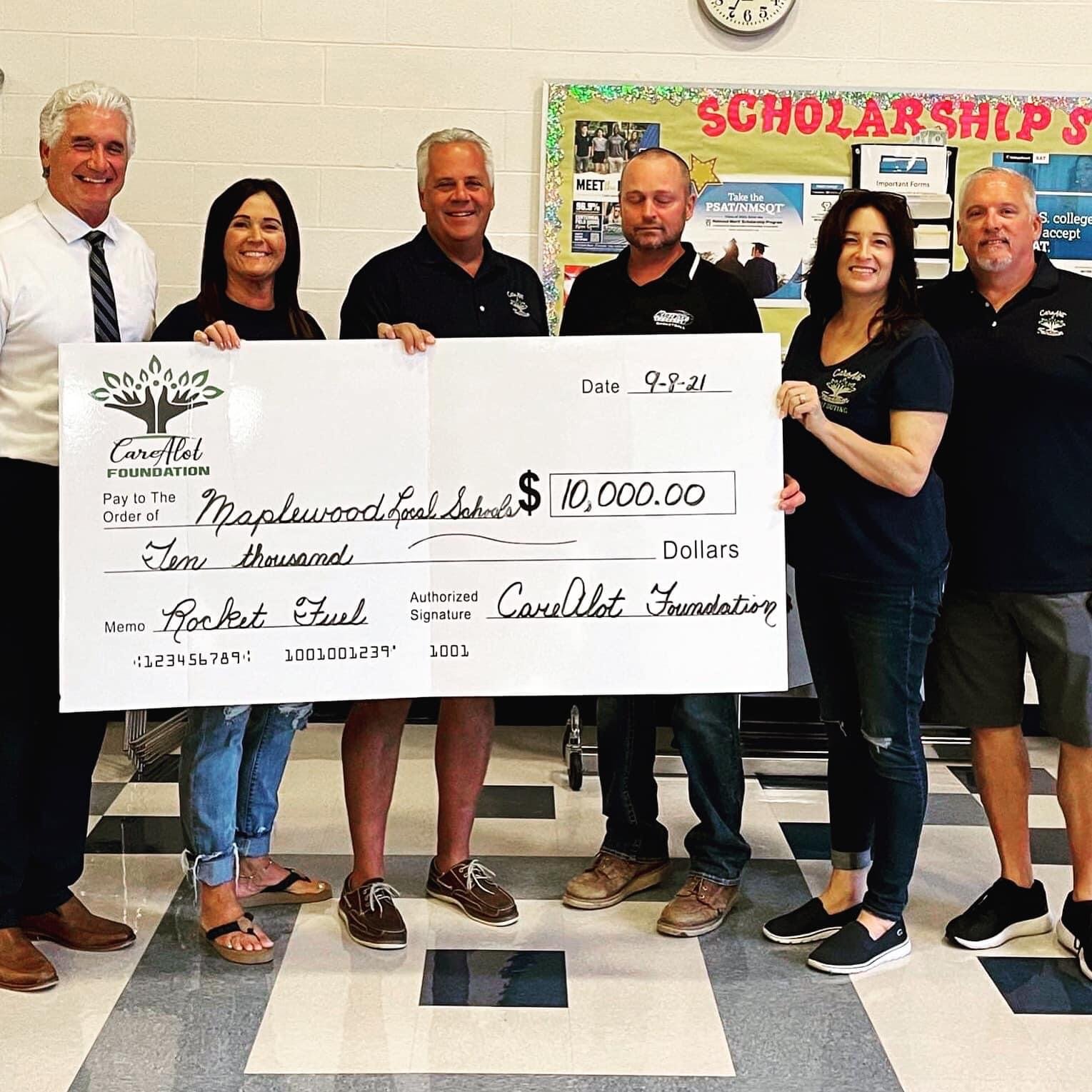 Group of seven people standing in a hallway, holding a large ceremonial check for $10,000 made out to Maplewood Local Schools, dated 9-8-21, from the Carelott Foundation. They are smiling and dressed in casual and business casual attire. Behind them 