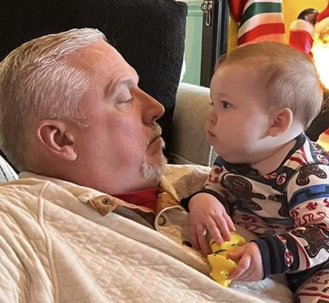 An older man with closed eyes lying on a couch, facing a young child who is leaning over him with their hands on the couch, both making eye contact. The man has light-colored hair and a mustache, and the child is dressed in pajamas with a patterned d