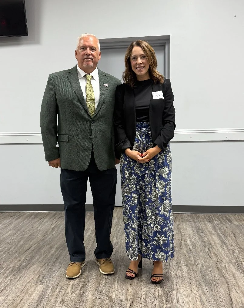 A man and a woman standing side by side indoors, dressed in business attire, smiling at the camera.