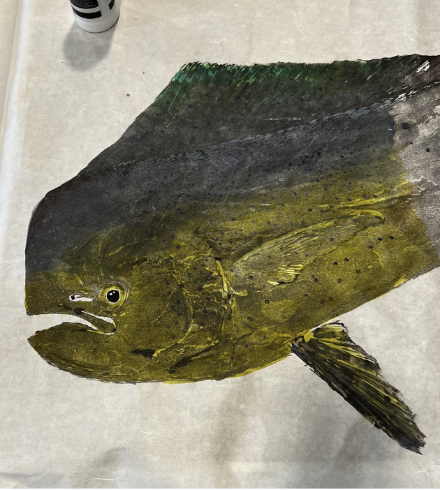 A realistic painting of a fish, possibly a yellow tang, on a light-colored surface with art supplies visible in the background.
