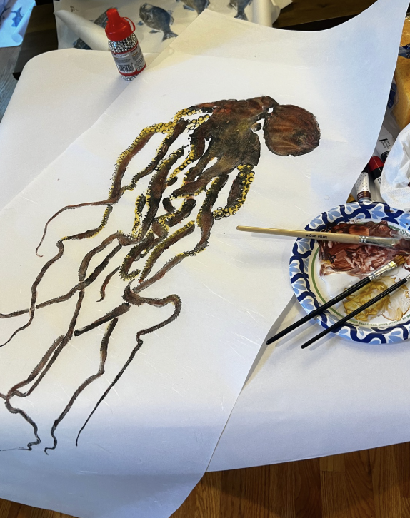An art project depicting an octopus with detailed tentacles on a large sheet of paper. Art supplies including brushes, paint, and a paper plate are visible on the table.