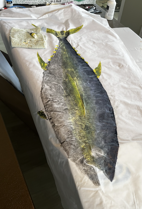 A large, detailed illustration of a fish painted on a cloth or canvas, with many tools and supplies around it, resembling a work in progress or artwork creation setup.