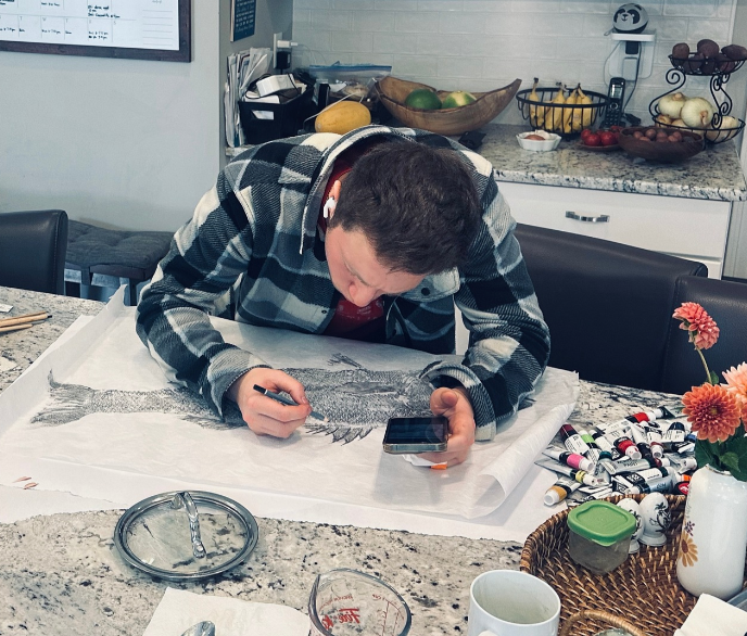 Person sketching a fish on a large sheet of paper while looking at their phone in a kitchen with fruit and flowers on the counter.