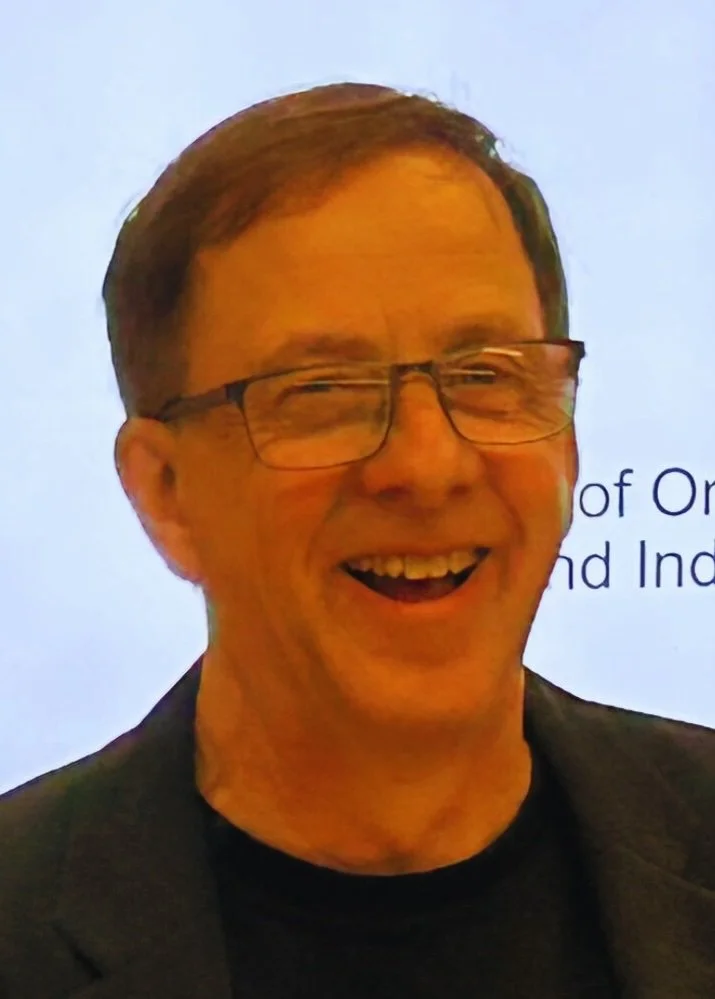 Smiling man with glasses, wearing a dark blazer and black shirt, with a light background showing partial text.