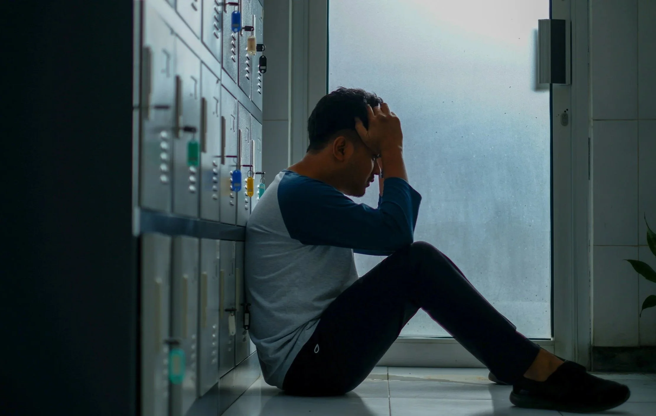 Photo of a man sitting on the floor next to a row of lockers with his head in his hands