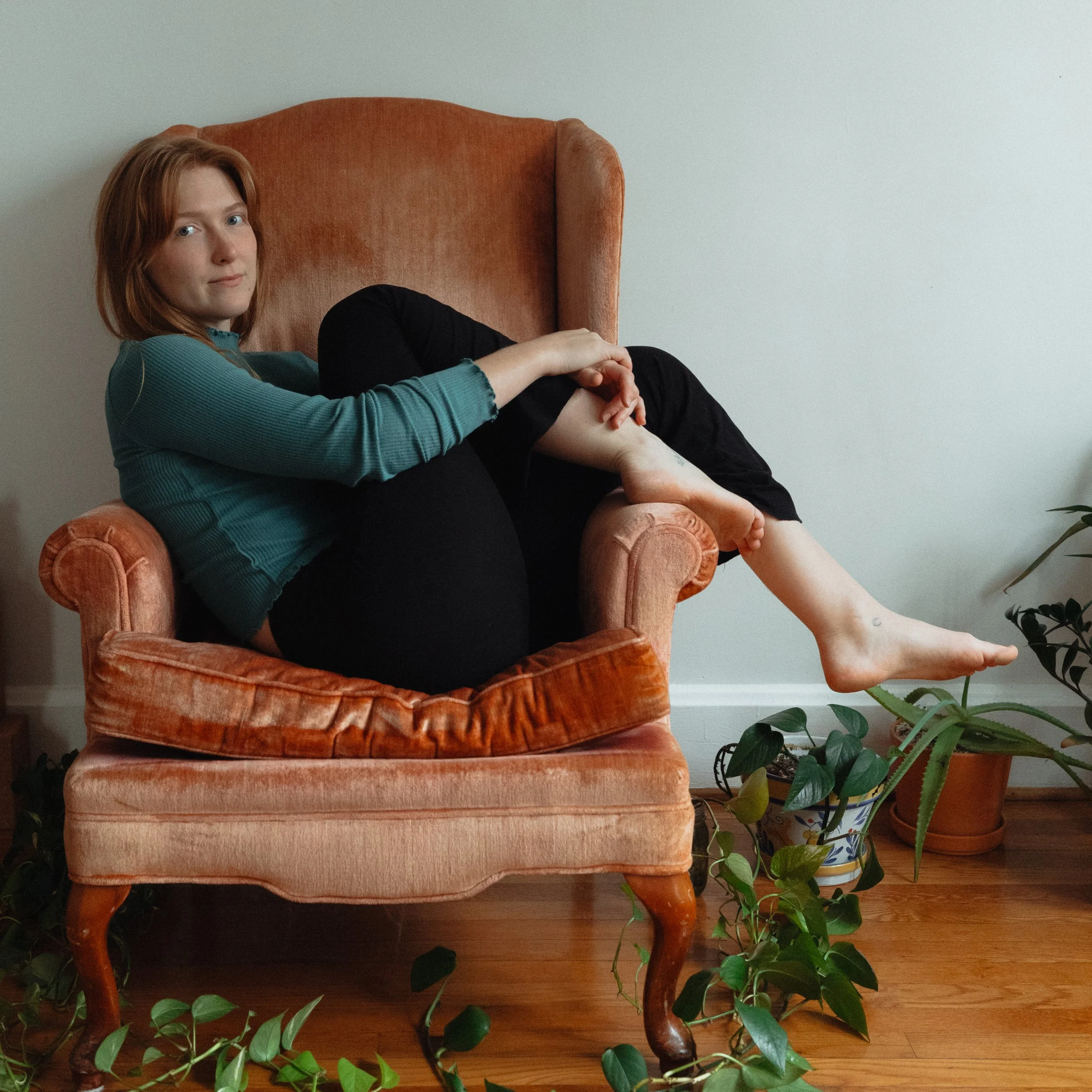 Young woman with red hair sitting on an orange vintage armchair, wearing a teal long sleeve top and black pants, with bare feet resting on a plant pot and the armrest of the chair. The scene is indoors with a white wall and wooden floor.