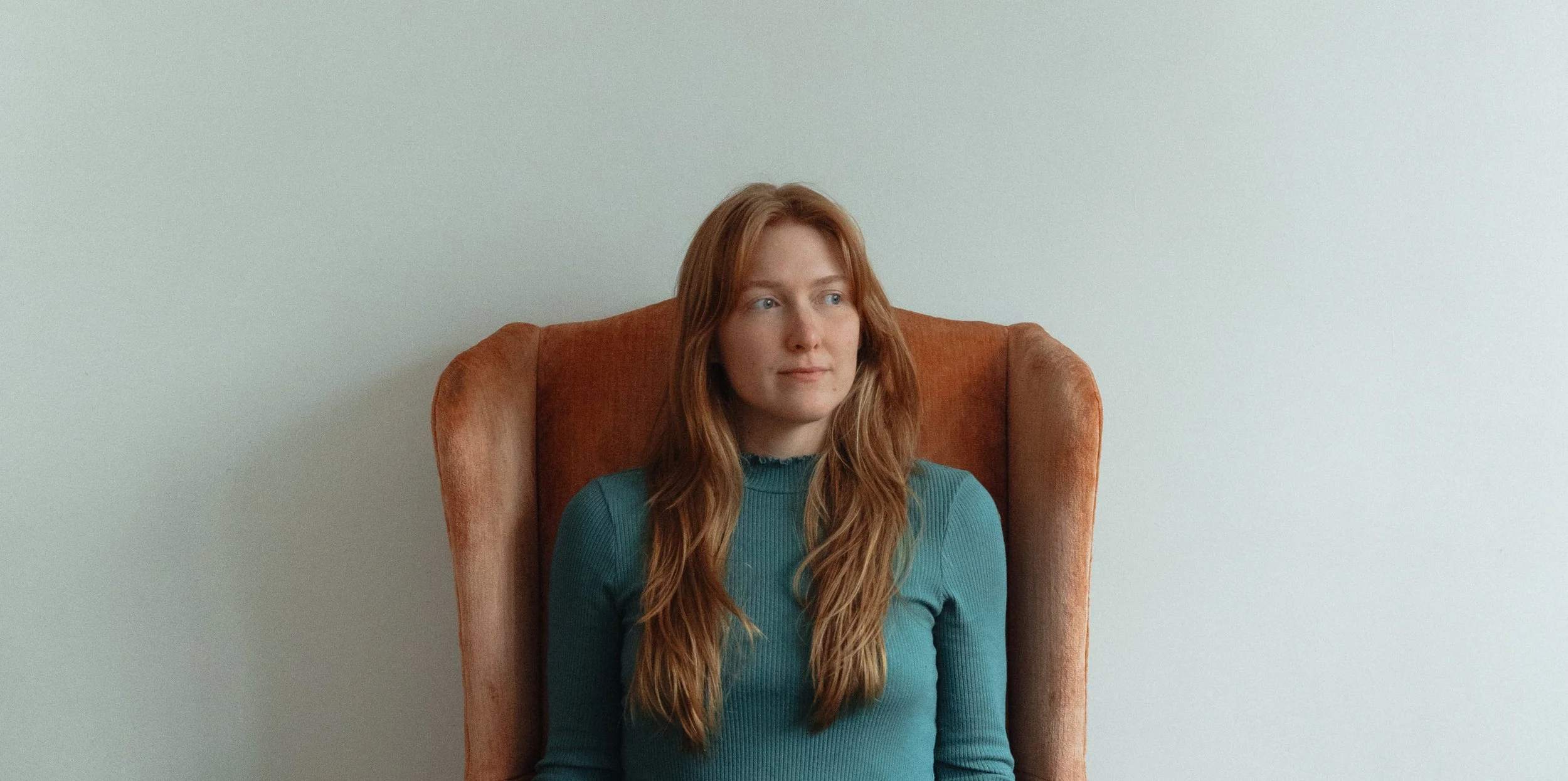 A woman with long red hair sitting on an orange-brown armchair against a plain light-colored wall.