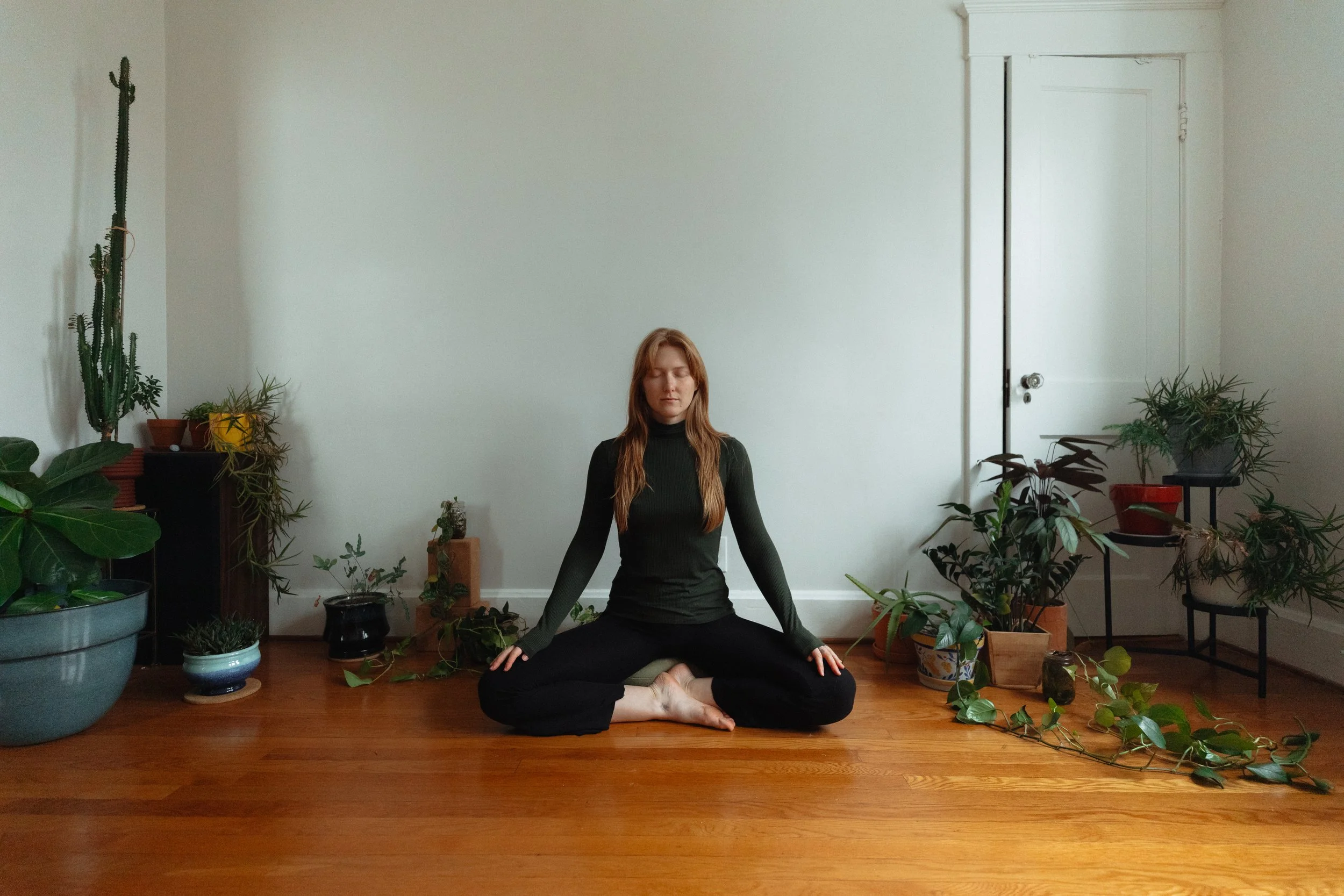 A woman with long red hair sitting cross-legged on a cushion in a room with hardwood floors and neutral walls. She is wearing a black long-sleeve top and black pants, with her eyes closed and hands resting on her knees. The room is decorated with numerous potted plants of various sizes on the floor and on stands.