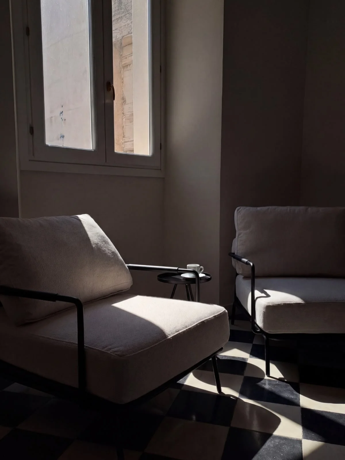The black-and-white floor is original to the house &mdash; the same checkerboard that has held footsteps here for generations. Now two seats sit on it, angled toward the window and the afternoon. Small signs that the rooms are nearly ready to be used