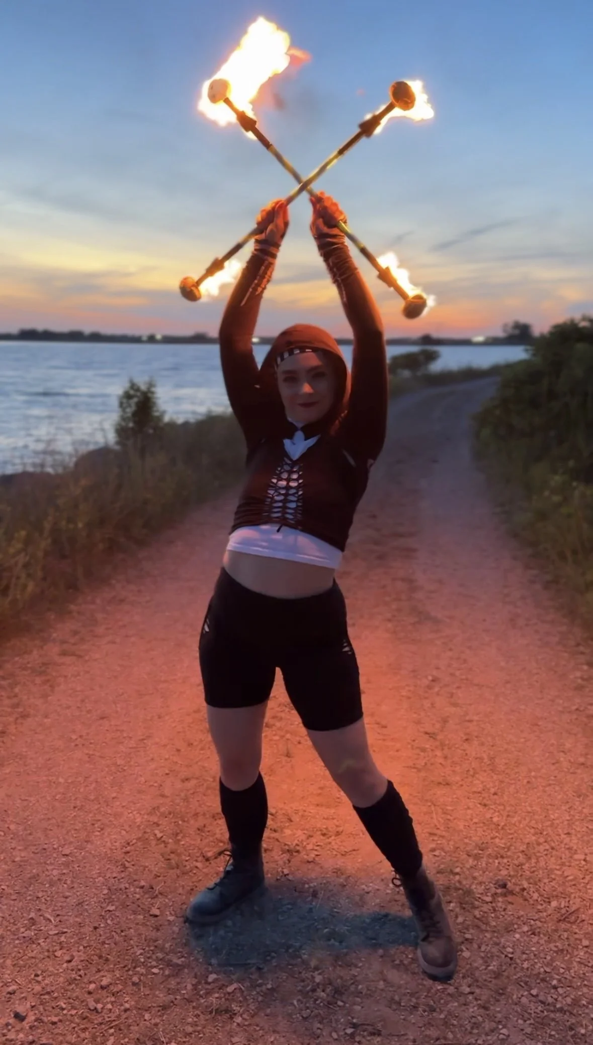 A person holding with both hands two fire-torches crossed above their head on a dirt path near a body of water at sunset.