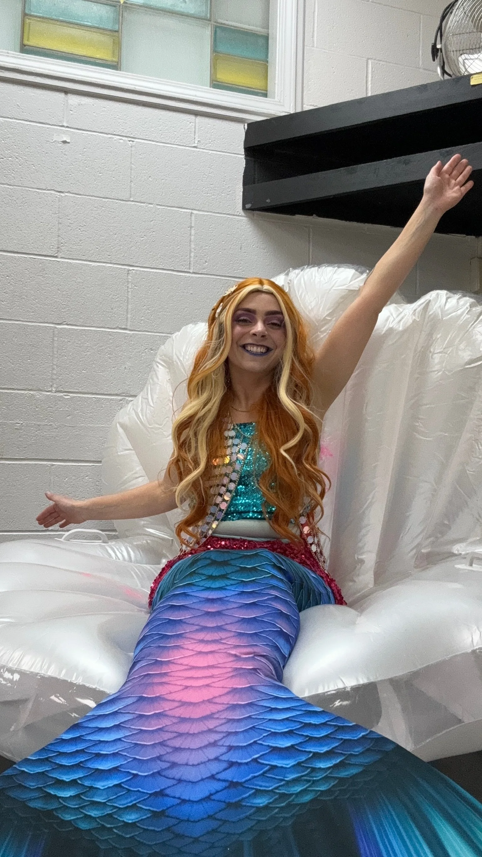 A woman dressed as a mermaid, sitting on a white inflatable float, smiling with her arms stretched out, in a room with white brick walls and a window with colorful stained glass.