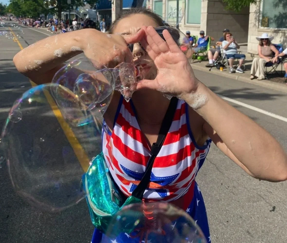 A woman making a heart shape with her hands as she blows bubbles during a parade or festival on a sunny day, with people sitting on the sidewalk in the background.