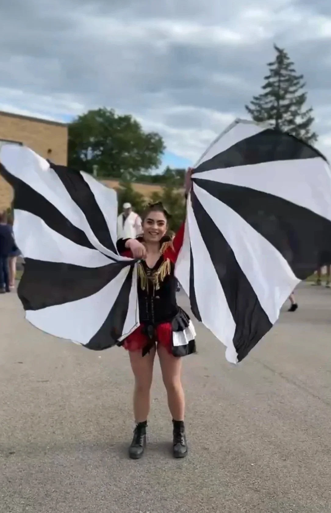 A woman dressed in a black, red, and gold costume holding large black and white striped flags outdoors with other people and trees in the background.