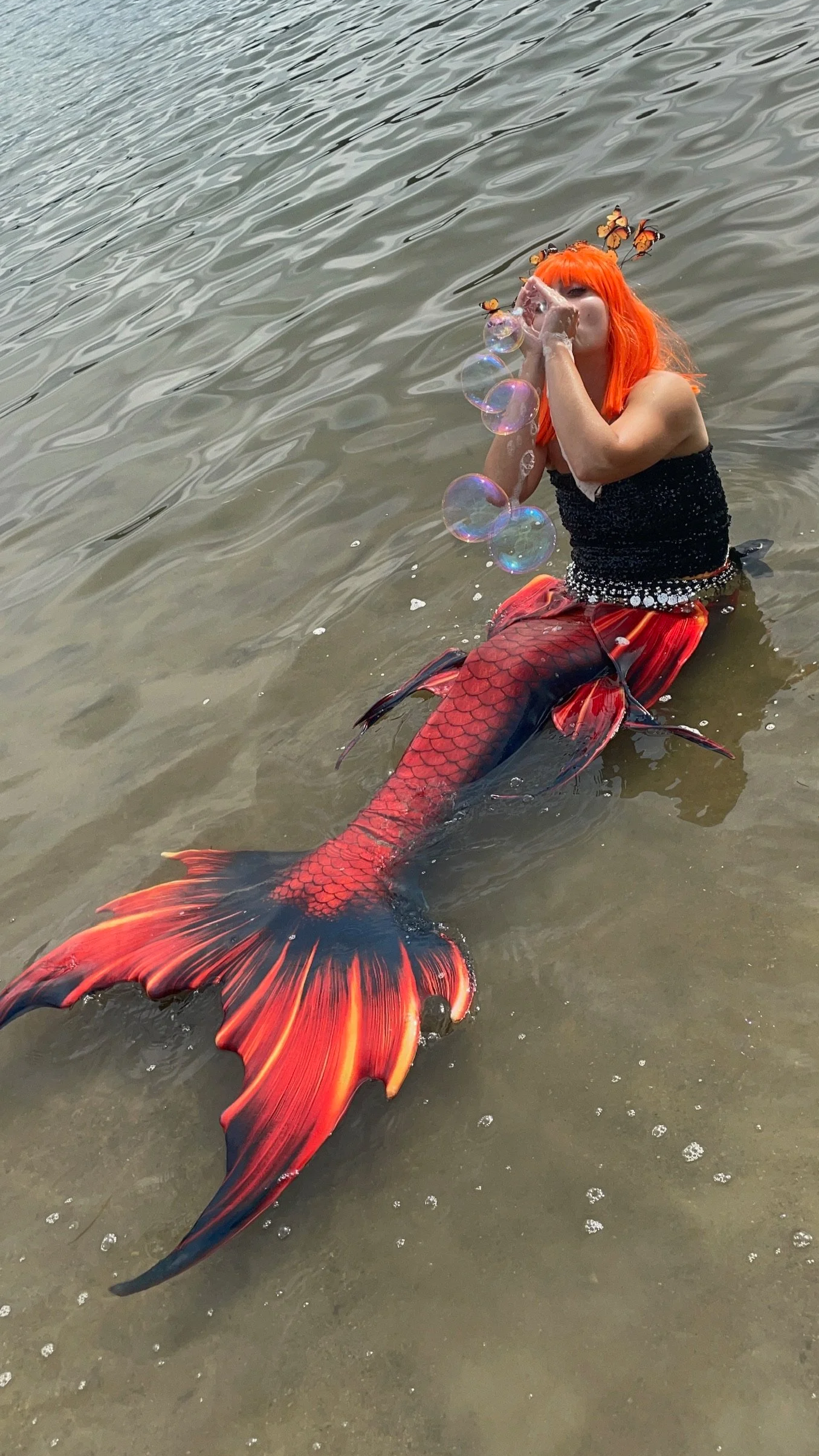 A person with bright orange hair wearing a black top and a red mermaid tail infused with scales, blowing bubbles while standing in the water.