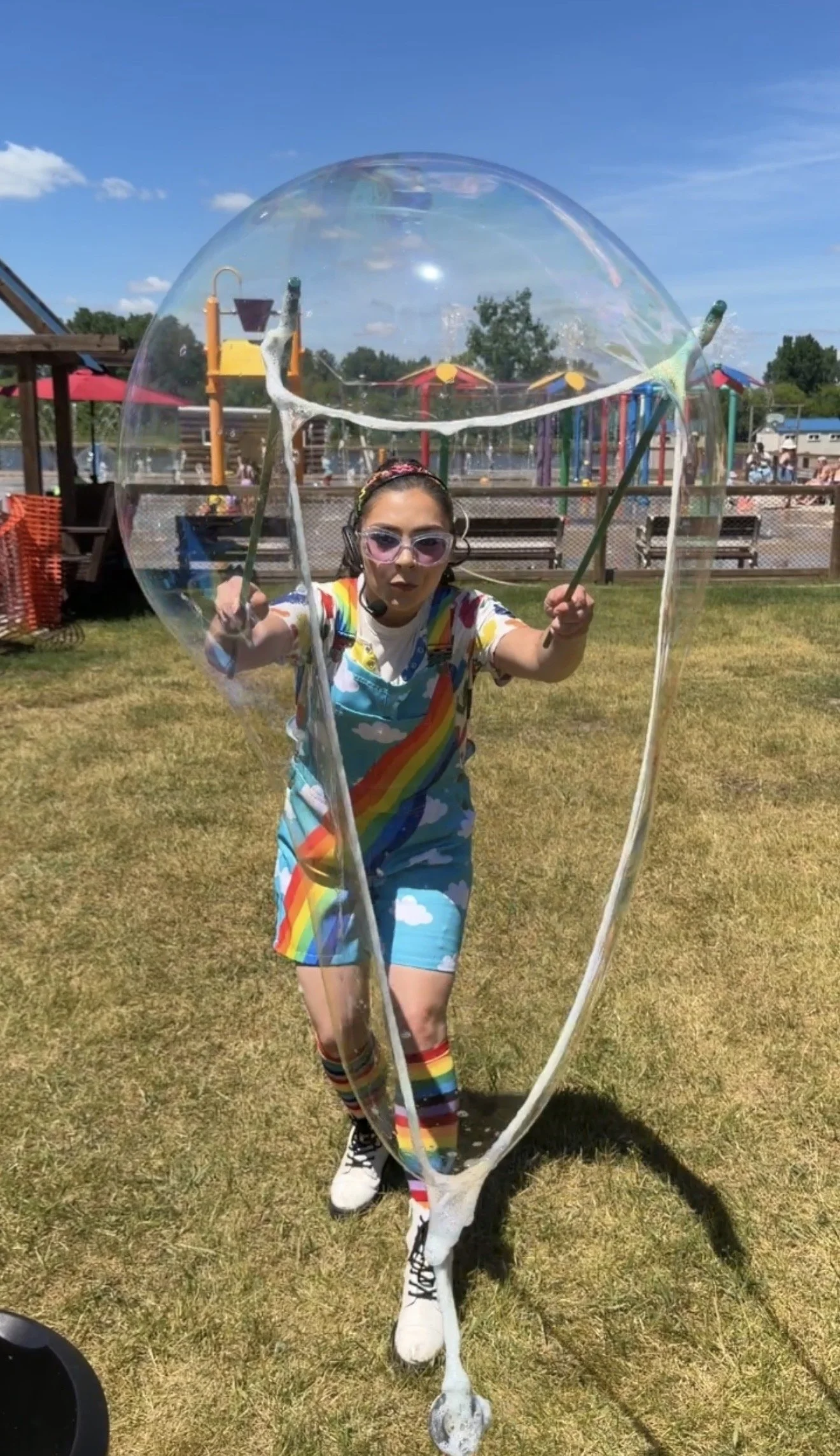 A young woman in a bright rainbow outfit blows a giant soap bubble into the camera