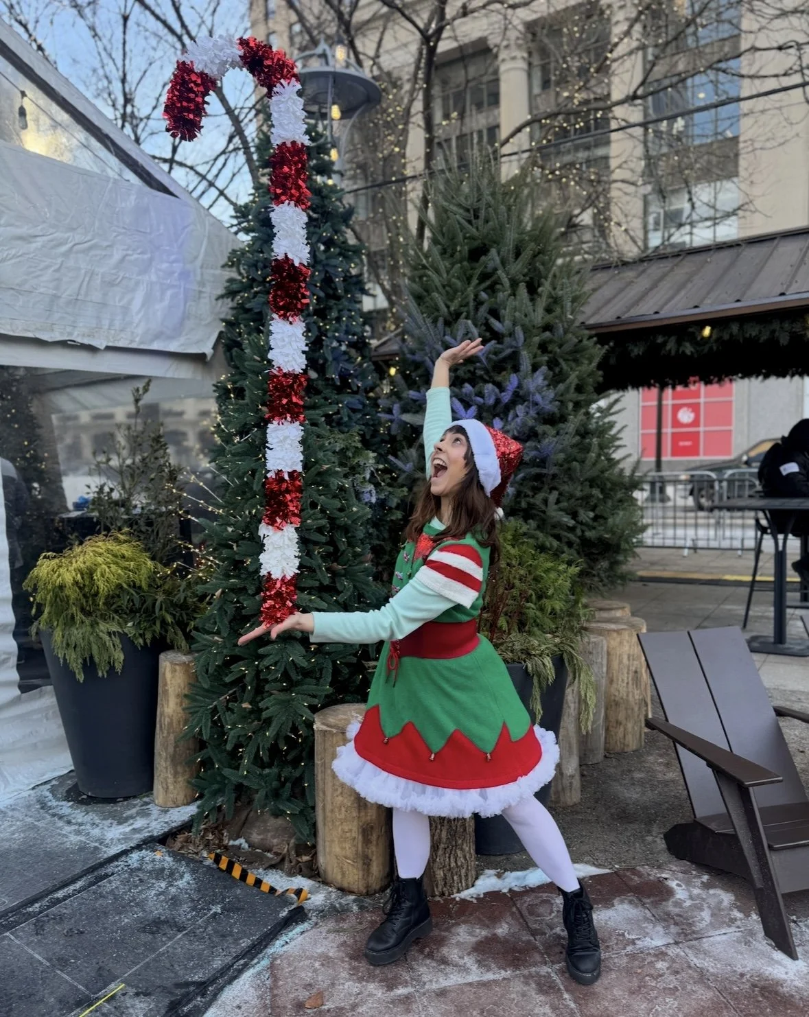 A woman dressed as an elf with a Christmas themed dress and Santa hat, standing outdoors near Christmas trees, smiling and reaching high.