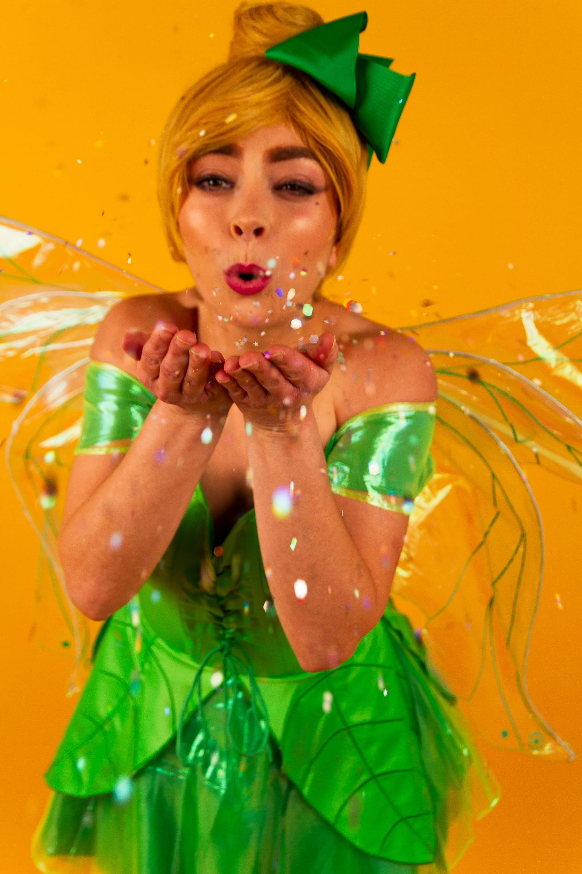 A woman in a bright green fairy costume with transparent wings and a large green bow in her hair blows confetti from her hands against a yellow background.