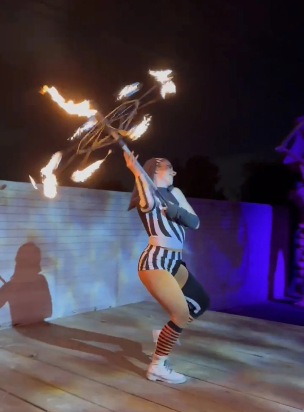 A female fire performer is spinning a flaming prop on a dark stage, dressed in a black and white striped outfit.