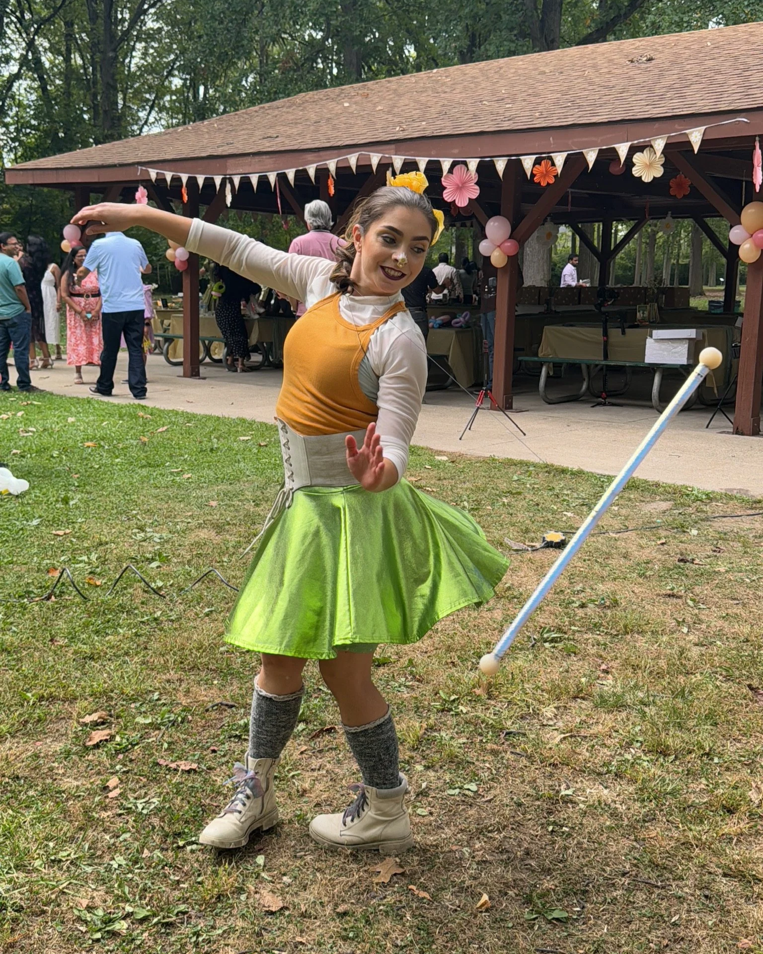 A woman dressed as a clown or performer twirling with a green skirt and a mallet in a park setting during an outdoor event, with people and a decorated pavilion in the background.