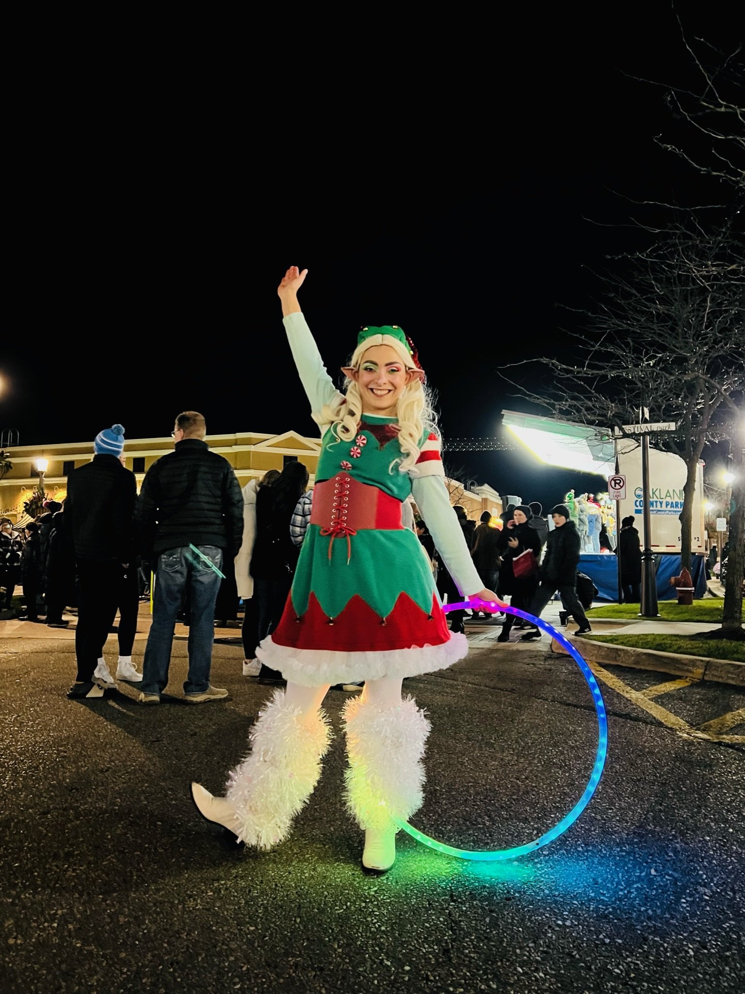 A smiling woman dressed as an elf with a green and red holiday costume, furry white boots, and holding a lit LED hoop, standing outdoors at night during a holiday event.