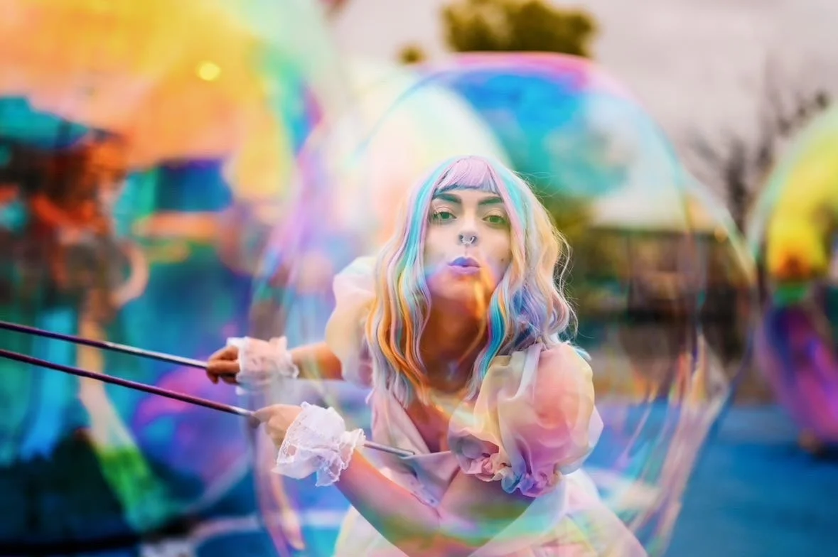 A young woman with rainbow-colored hair blowing a large, colorful bubble outdoors during sunset.