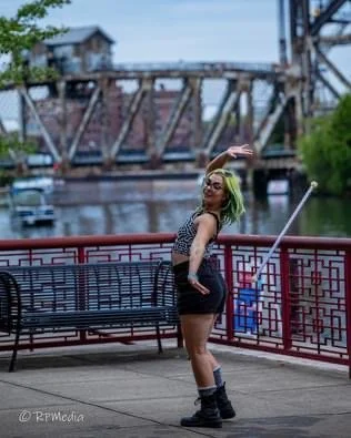 A woman with green hair posing on a bridge near a river, with trees and a building in the background.