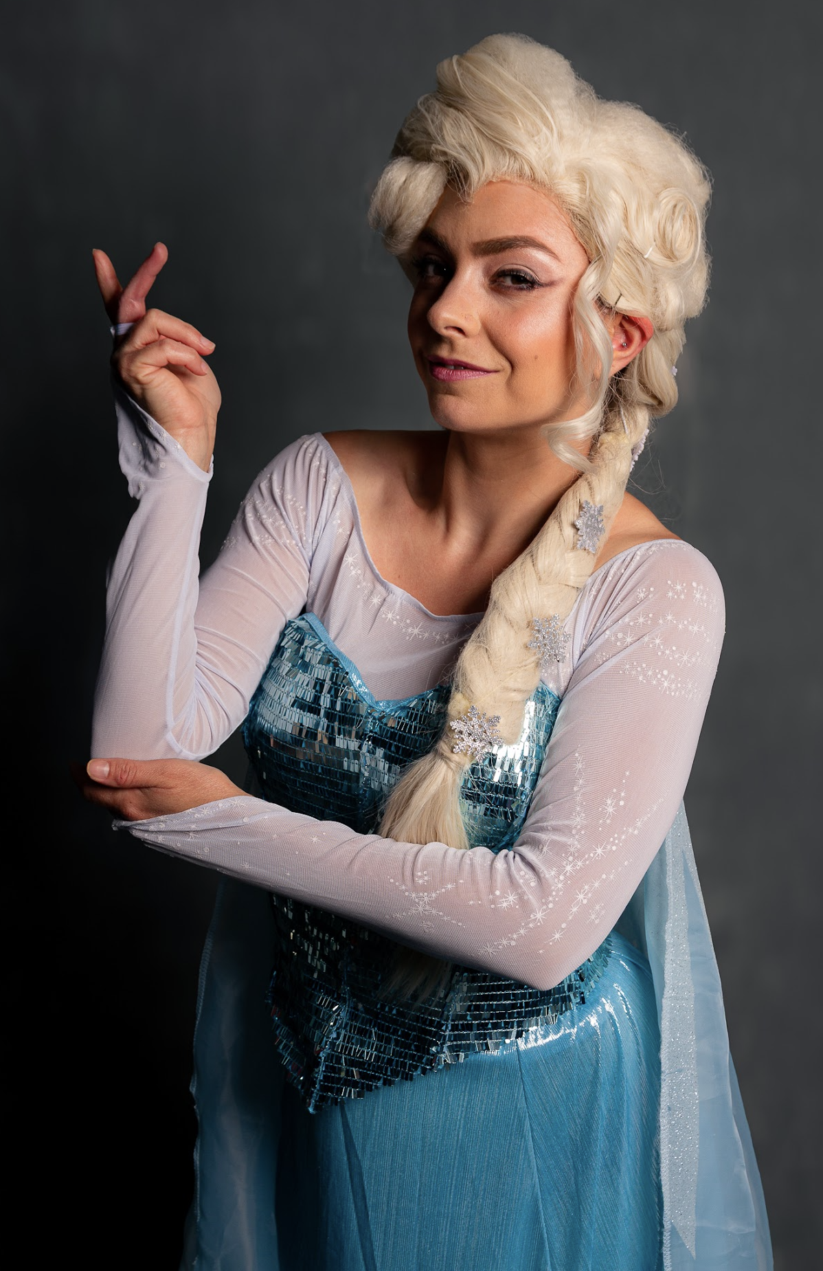 Woman dressed as Elsa from Frozen, with platinum blonde hair in a braid, wearing a blue sequined dress with sheer sleeves and snowflake accents, posing with a confident expression
