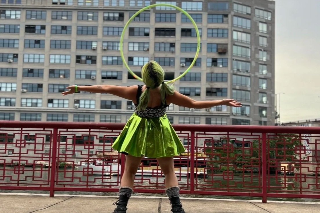 A person with green hair and a green metallic dress standing on a balcony, with arms outstretched and a large yellow hula hoop behind their head.