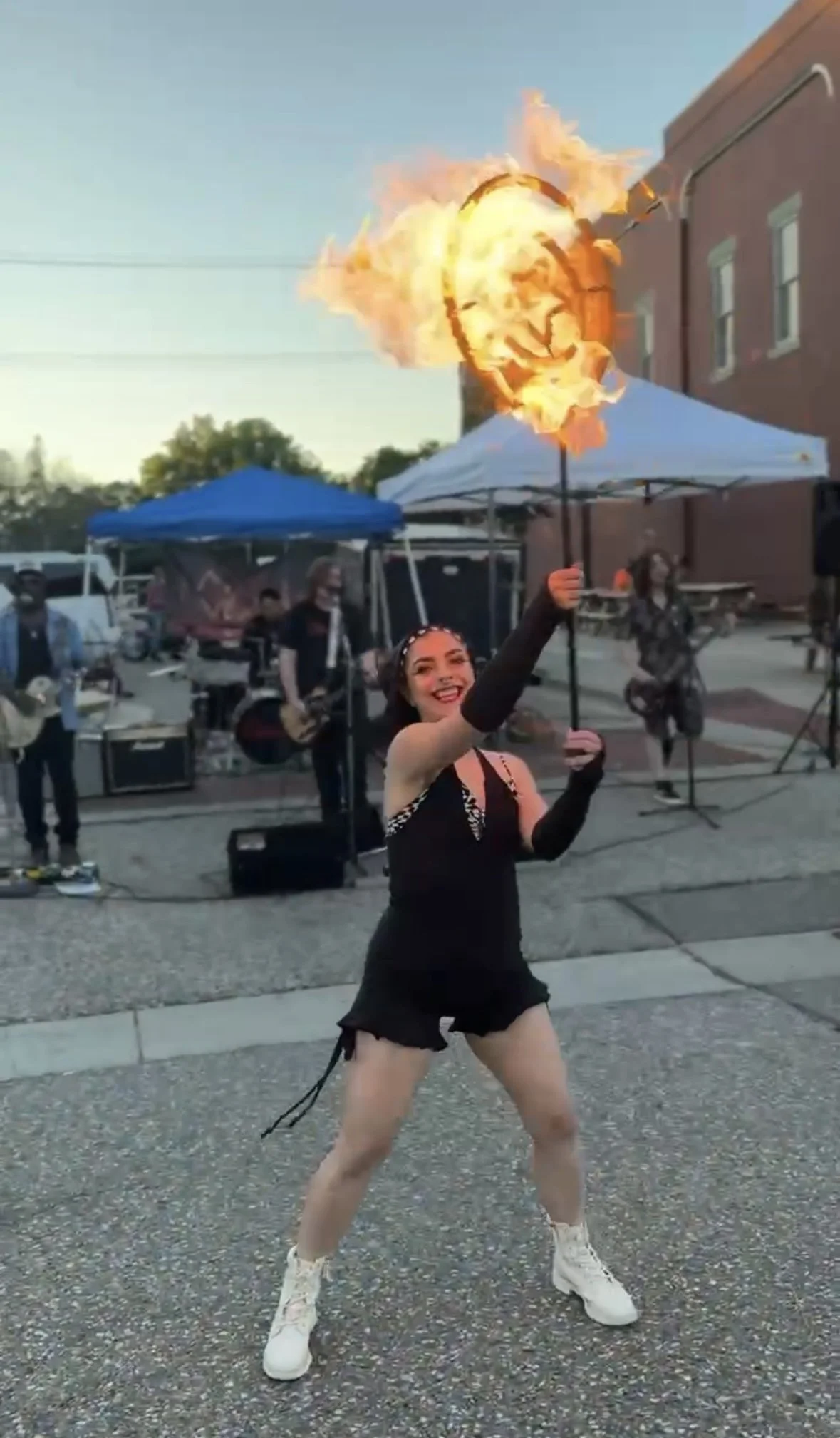 A woman performing a fire act outdoors, holding a flaming torch in the air during a street performance with a band playing in the background.