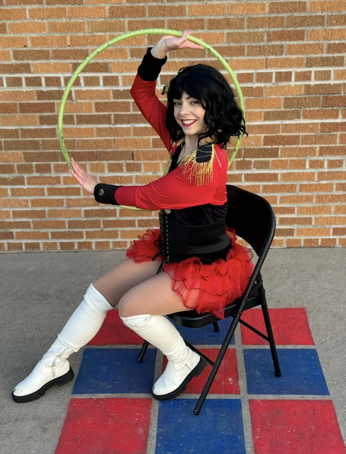 A woman dressed as a circus performer or clown sitting on a black chair on a red and blue checkered mat, holding a green hoop, with a brick wall background.