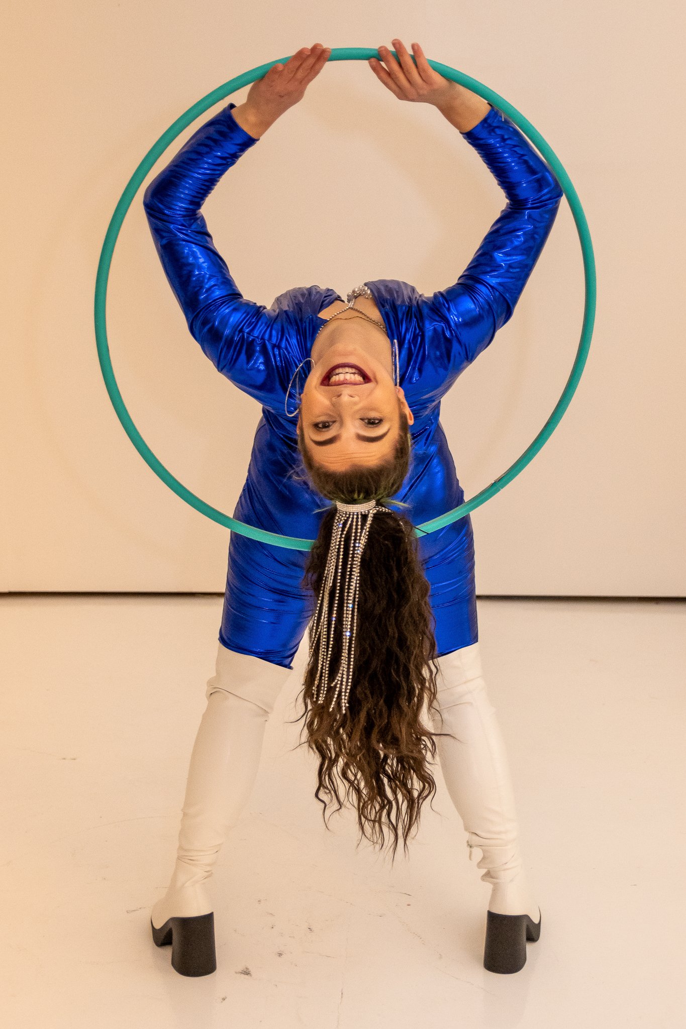 Woman in a blue shiny outfit performing an acrobatic pose with a green hula hoop, standing on white platform shoes, with long, curly hair and pearl accessories.