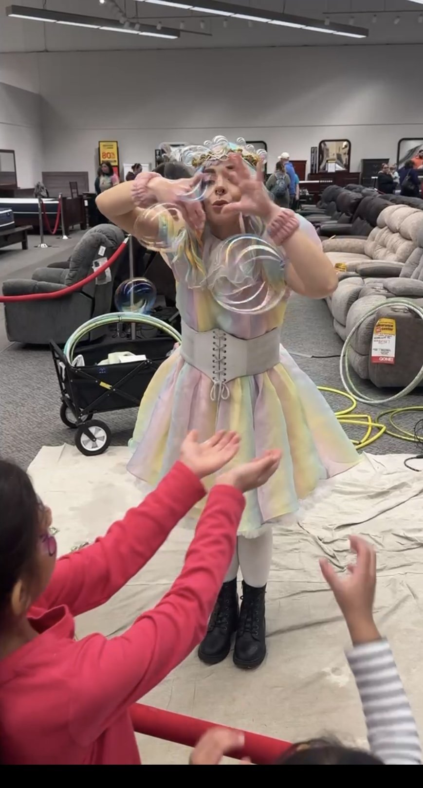 A woman dressed in a pastel-colored dress with a wide belt is making large soap bubbles at an indoor event, with children reaching out to catch or pop the bubbles, in what appears to be a furniture store.