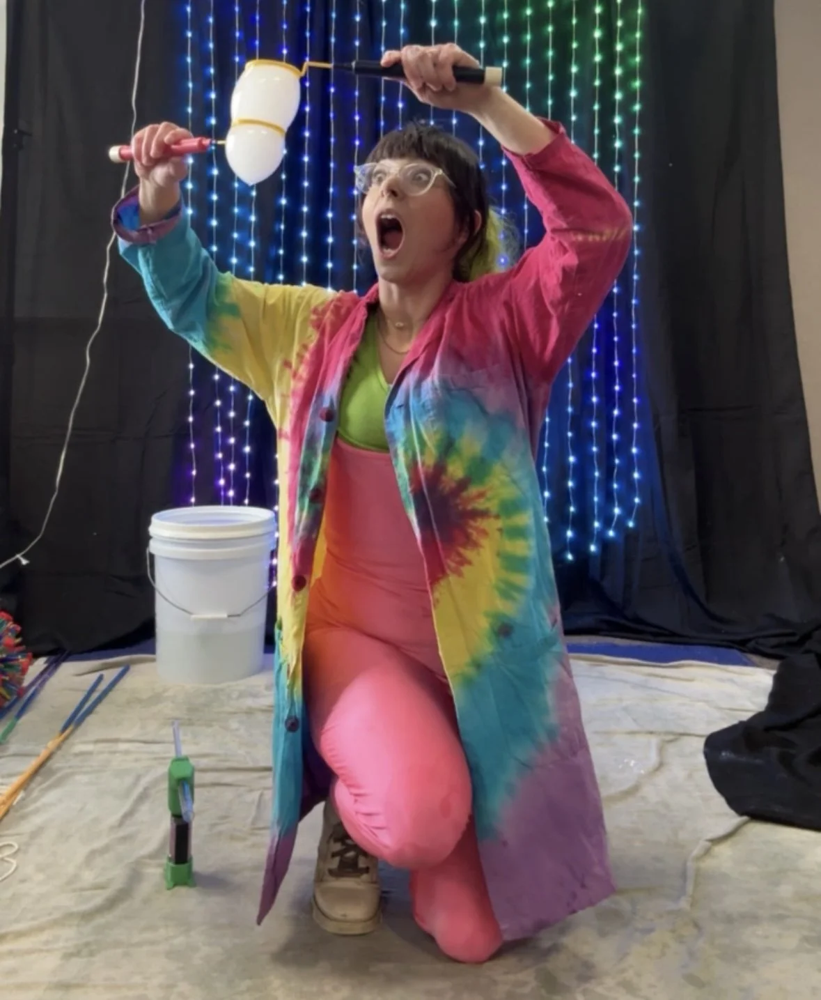 Person in a colorful tie-dye coat and pink clothing with glasses, holding a mallet above their head, appearing to strike a balloon. The background has blue and green string lights, and there are pick-up sticks and a bucket on the floor.