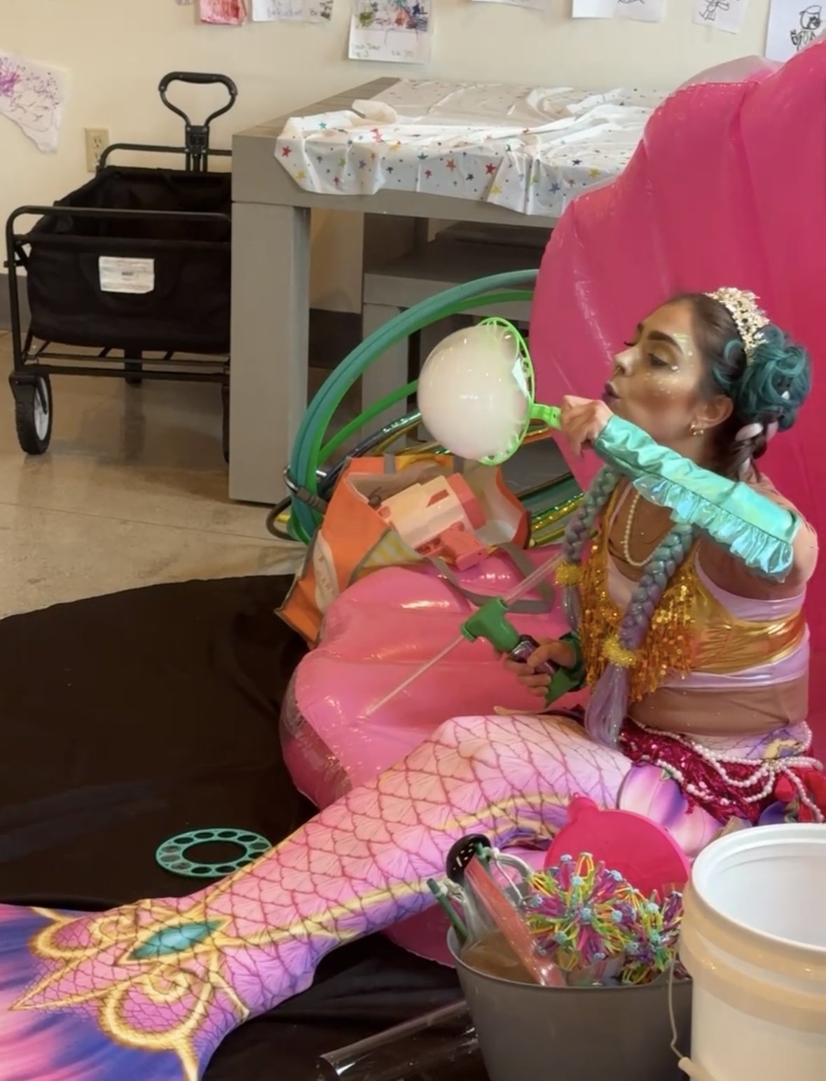 A woman dressed in colorful, fairy-tale-like costume with a tiara and makeup, sitting on the floor, blowing bubbles from a bubble wand.