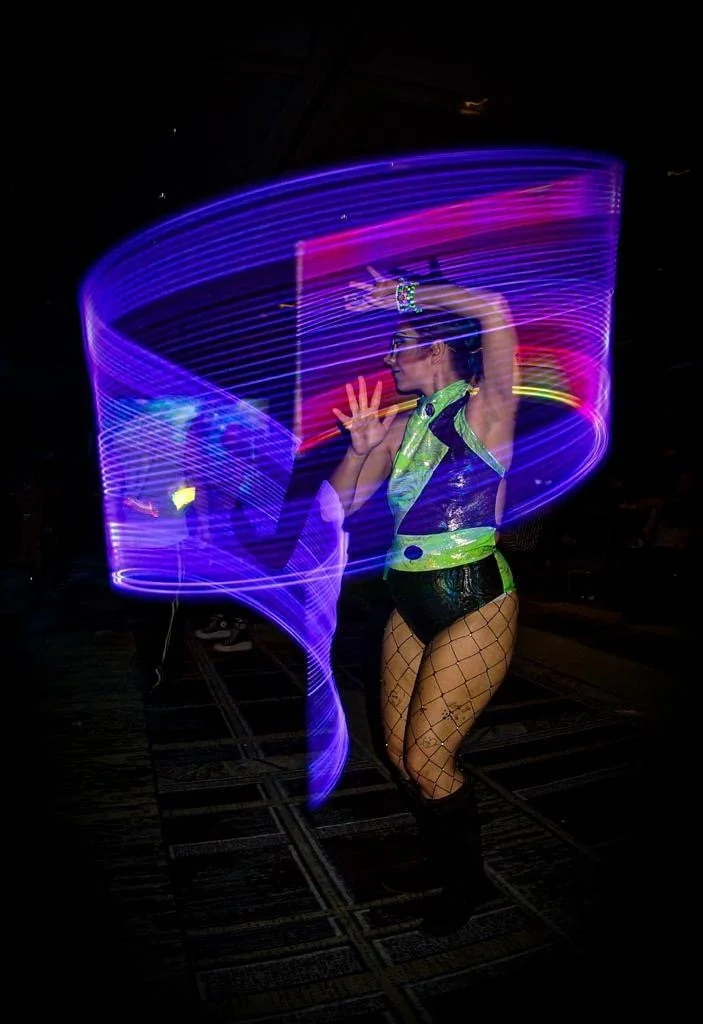 A woman performing a dance with colorful LED lights creating swirling light trails around her in a dark environment.