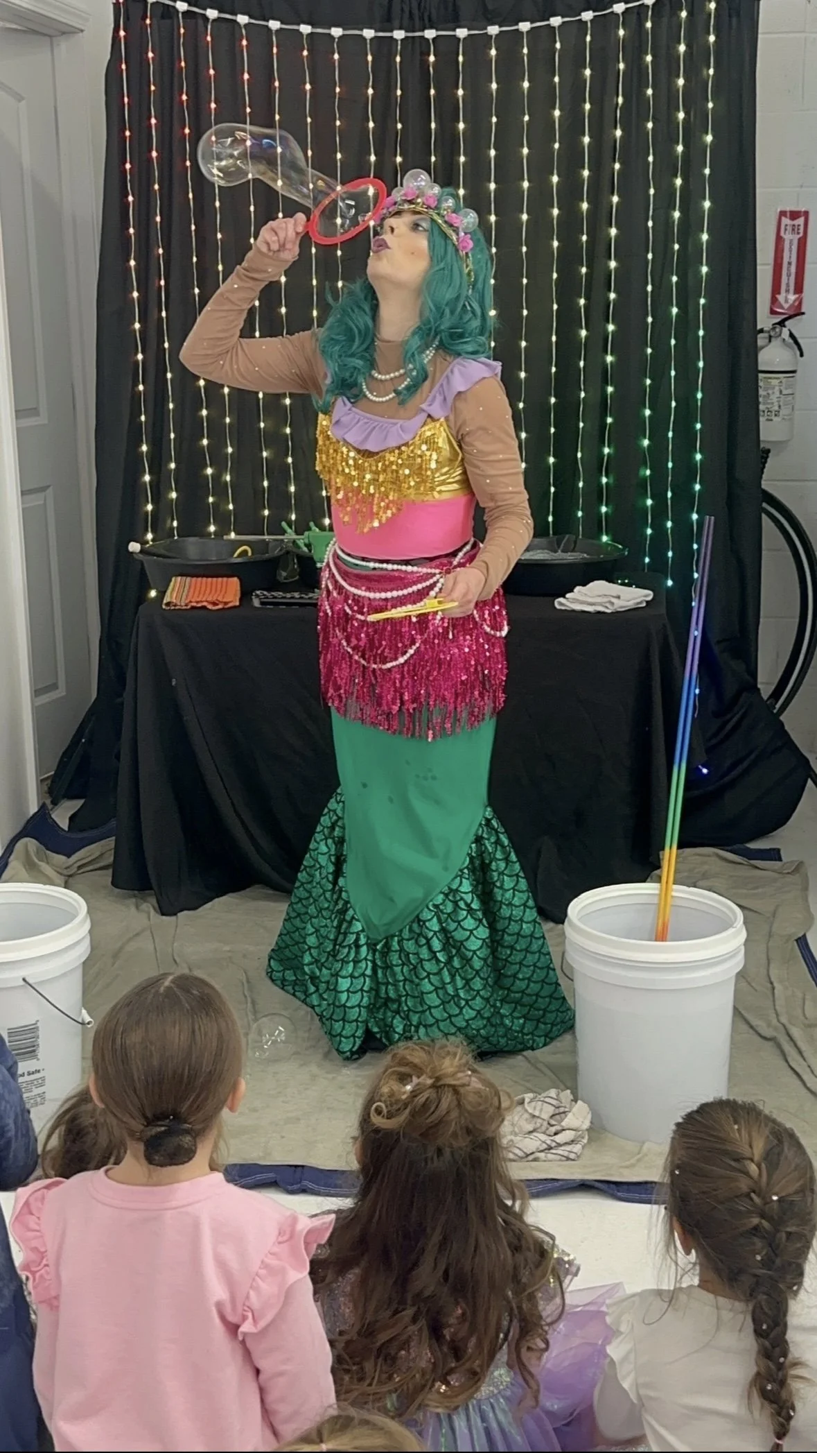 A woman dressed as a mermaid with teal hair and a floral headband is blowing a large soap bubble at a children's event, with children watching in front of her.