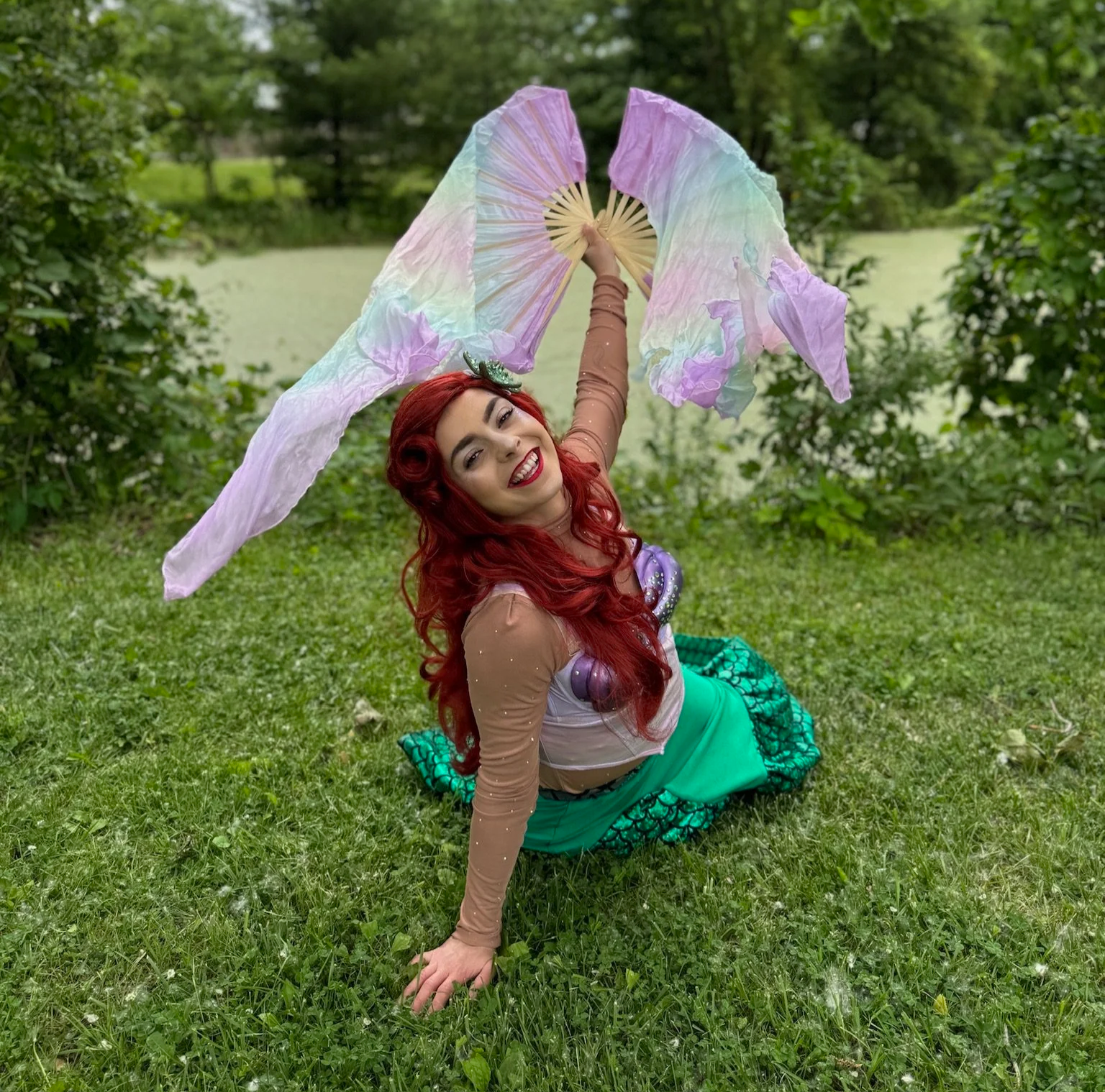 A woman dressed as a mermaid with long red hair, wearing a sparkly top and green tail, poses outdoors on grass with a pond and trees in the background. She is holding iridescent purple and pastel-colored fabric in the air and smiling.